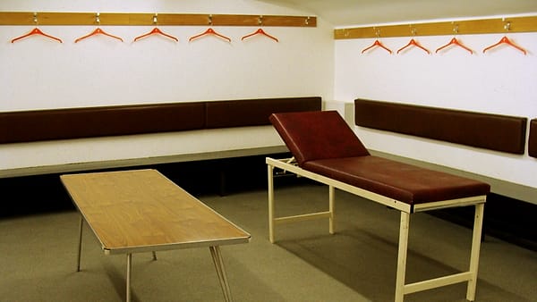 A quiet treatment room with a covered massage table, folded towels on a shelf and a low lamp beside a closed door