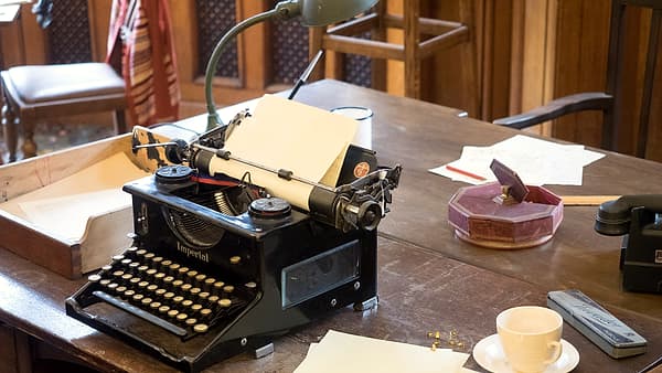 A laptop and a printed form share a small desk, with a coffee ring on the paper and a pen lying across the keyboard
