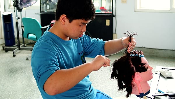 Students at a training salon practicing haircutting on mannequin heads clamped to a row of workstations