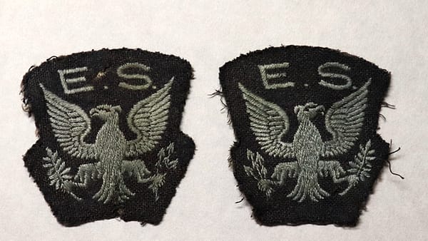 A plain navy uniform shirt on a hanger with an unbranded shoulder patch and empty epaulettes against a locker door