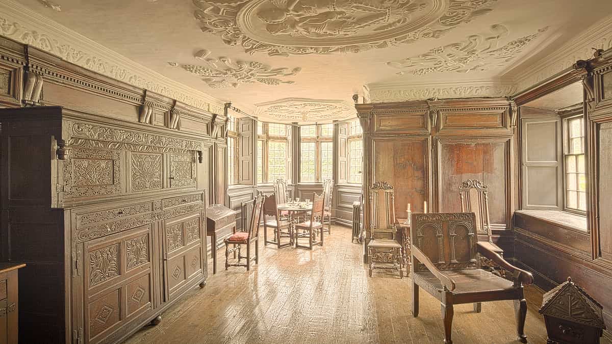 An empty wooden chair beside a low railing in a quiet paneled room with daylight from a high window